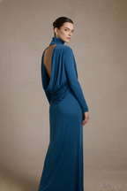 A woman modeling a floor-length teal blue gown from the back, featuring a draped open back, long sleeves, and a high neck, set against a textured beige studio backdrop.